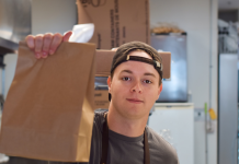 A young man with his hat on backwards, holding up a to-go brown bag