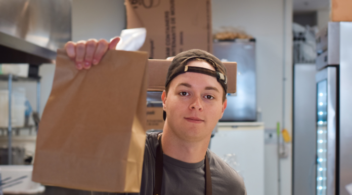 A young man with his hat on backwards, holding up a to-go brown bag
